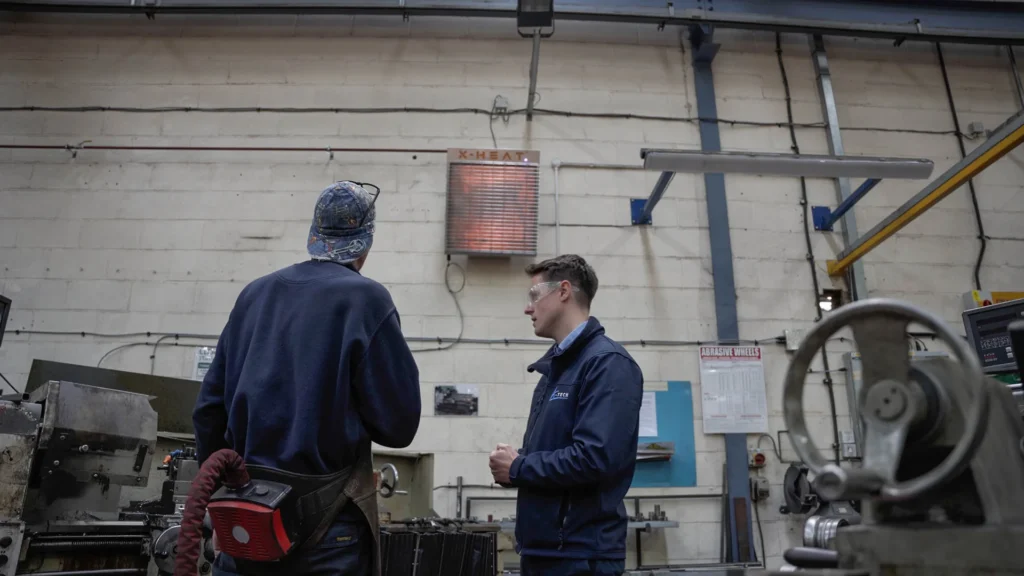 two workers standing underneath a x-heat heater that is turned on.